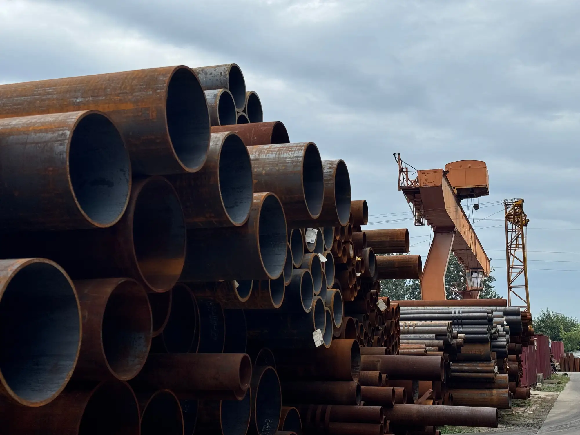 Stacks of steel pipe in an open storage yard with a gantry crane behind